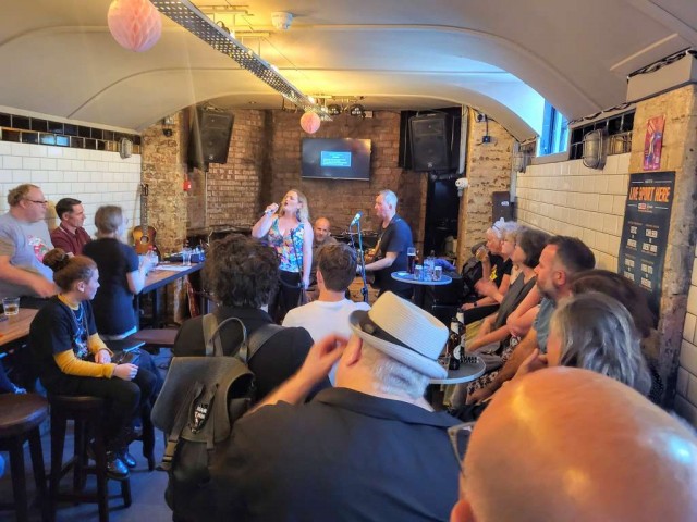 Pub lounge full of people as a woman sings at the front with man playing guitar.